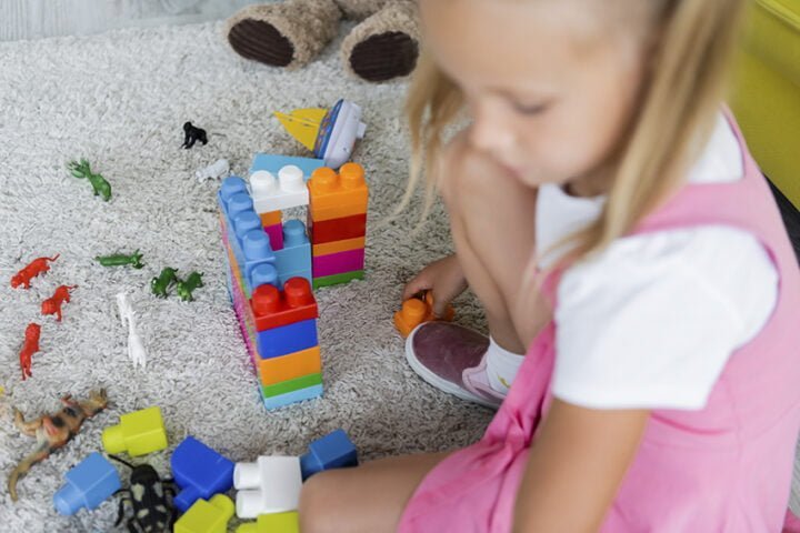 blurred girl in pink dress playing building blocks on carpet in kindergarten playroom